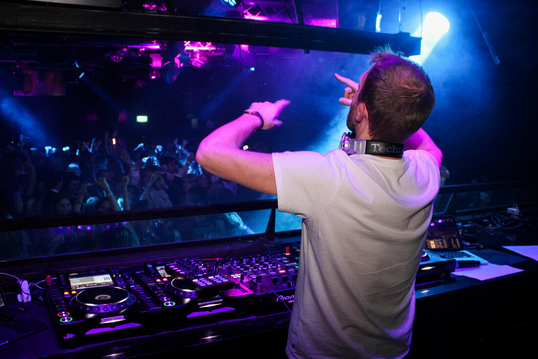 A DJ in a white T-shirt at the decks, playing to a dark club lit blue